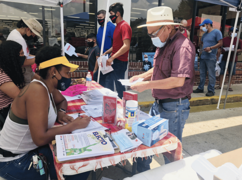 An outreach table staffed by Alabama Coalition of Immigrant Justice volunteers, interacting with a masked person, providing information on COVID vaccine equity along with masks, hand sanitizer, and more personal protection equipment.