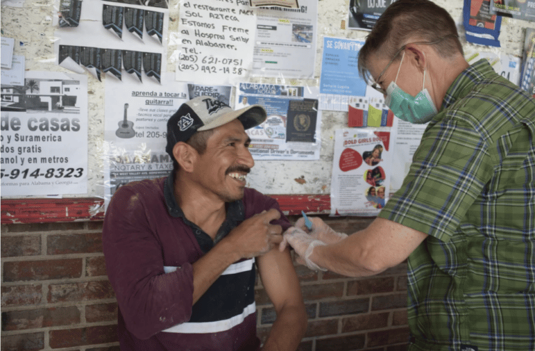 A smiling Latinx man wearing a cap lifts his sleeve for a masked volunteer worker to administer a COVID vaccine