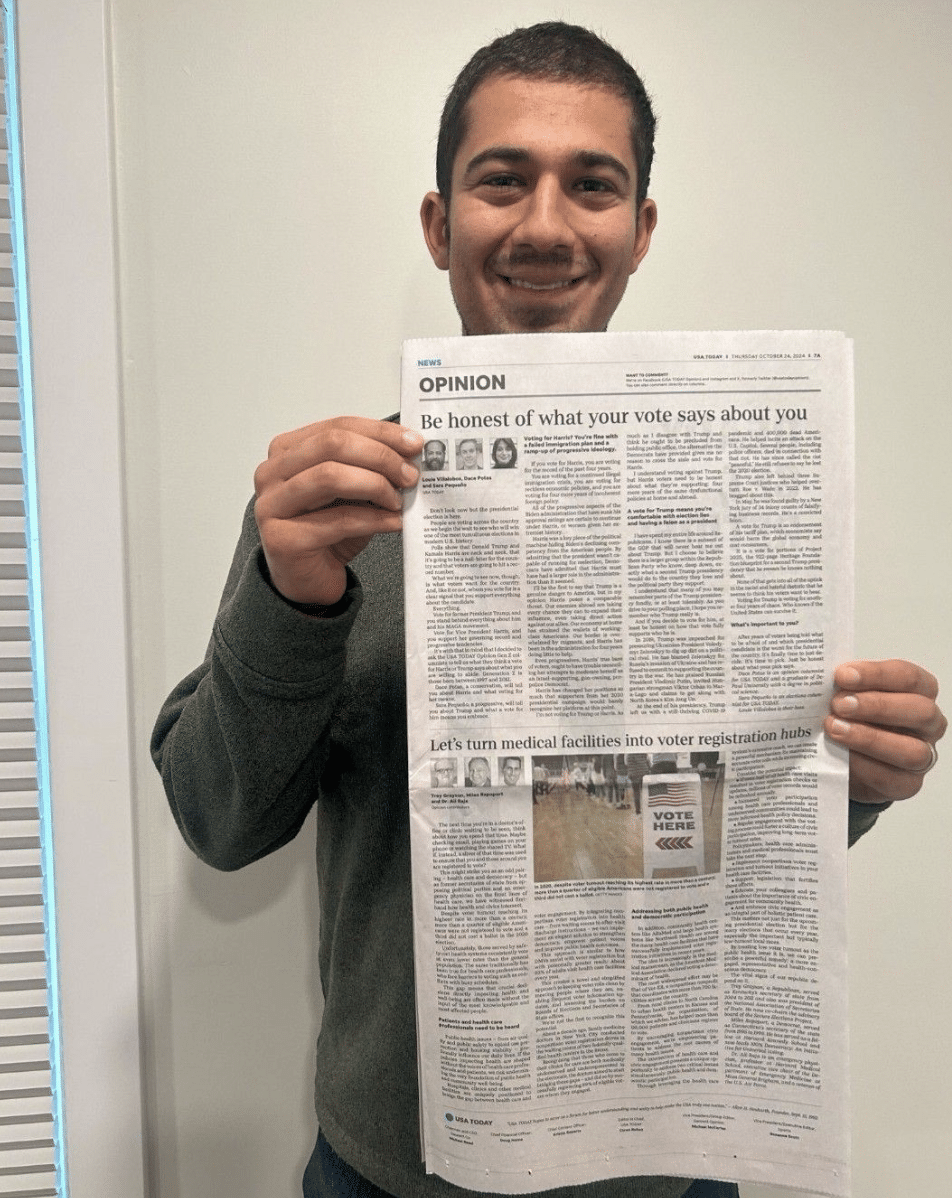 A man, smiling, holds up a print issue of an op-ed in USA Today